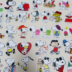 Peut inclure: Une collection d'autocollants Snoopy dans diverses poses et tenues. Les autocollants présentent Snoopy dans différentes situations, notamment portant un chapeau de pilote, tenant une bougie et avec un cœur. Les autocollants sont dans une variété de couleurs, dont le rouge, le jaune et le bleu.