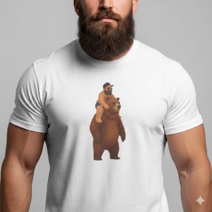 May include: White t-shirt featuring a cartoon illustration of a man riding a brown bear. The man is wearing a black cap and blue shorts. The bear is standing upright. The shirt is made of a soft, white material.