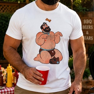 Puede incluir: Una camiseta blanca de manga corta con una ilustración gráfica de un personaje barbudo con collar y calzoncillos marrones, arrodillado mientras intenta alcanzar una golosina para perro, con una máscara de perro en el suelo cerca de las rodillas.