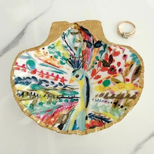 May include: A decorative scallop shell trinket dish featuring a hand-painted peacock design in vibrant shades of blue, yellow, red, and green with gold-painted edges, accompanied by a thin gold-toned metal ring with a small clear stone setting.