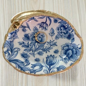 May include: A decorative seashell ring dish with a gold rim. The interior features a blue and white floral pattern, and a gold ring rests inside. The dish is set on a light wood surface.