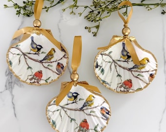 Può includere: Tre ornamenti a forma di conchiglia di capasanta bianca con illustrazioni colorate di vari uccelli appollaiati su rami marroni. Ogni conchiglia è bordata con vernice dorata e sormontata da una perlina dorata e un nastro di raso dorato.
