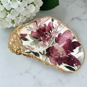 May include: A decorative oyster shell with a gold-painted edge. The shell is adorned with a floral design featuring burgundy and gray flowers on a white background. The shell is approximately 5 inches long.