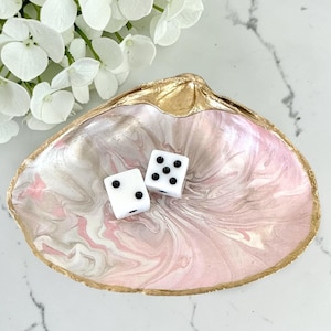 May include: A decorative seashell-shaped dish with a gold-painted rim and a pink and white marbled interior. Two white dice with black dots rest inside the dish. White flowers are in the background.