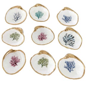 May include: A collection of nine white clam shells, each featuring a unique hand-painted coral illustration in shades of blue, green, or pink. The edges of every shell are finished with a metallic gold trim, creating a decorative set of small dishes.