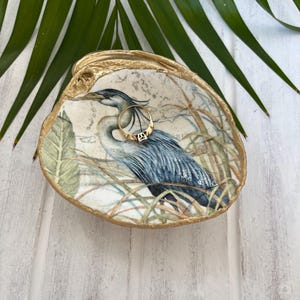 Pu&ograve; includere: Un piattino decorativo a forma di conchiglia con un'illustrazione dettagliata di un airone azzurro tra l'erba alta. I bordi della conchiglia sono dipinti con una finitura oro metallizzato. Un semplice anello d'oro riposa all'interno.