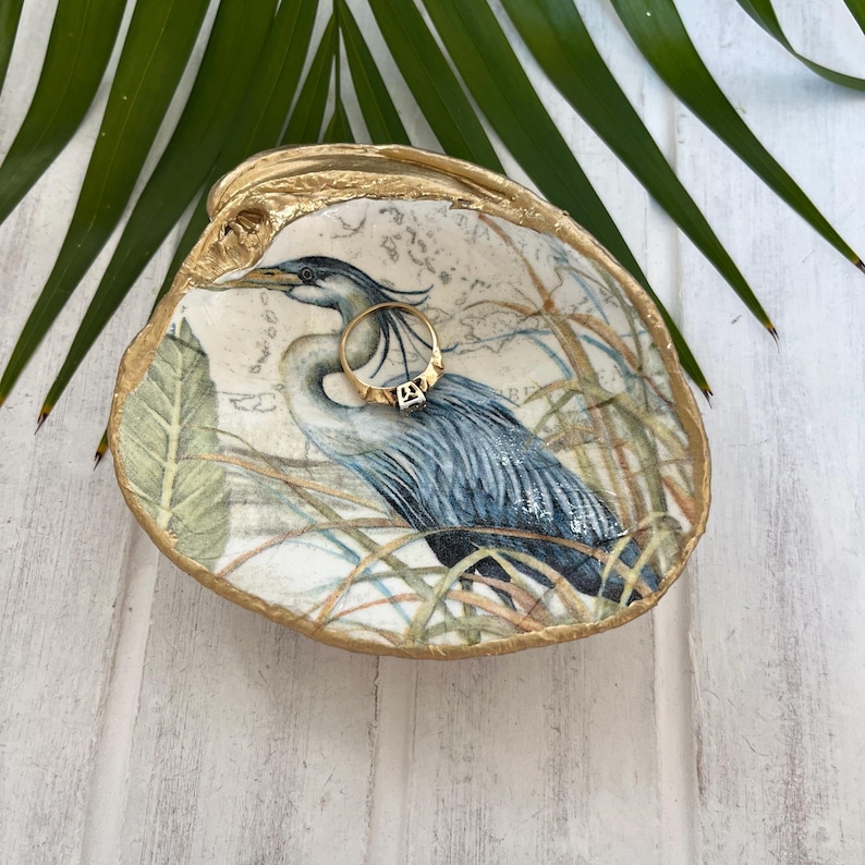 Pu&ograve; includere: Un piattino decorativo a forma di conchiglia con un'illustrazione dettagliata di un airone azzurro tra l'erba alta. I bordi della conchiglia sono dipinti con una finitura oro metallizzato. Un semplice anello d'oro riposa all'interno.