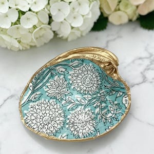 May include: A decorative seashell trinket dish with a gold-painted rim. The interior features a teal background with a white floral pattern. The dish is ideal for holding small jewelry items.