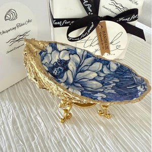 May include: A decorative oyster shell dish featuring a blue and white floral pattern on the interior and a gold-painted exterior edge. The shell is supported by a small, ornate gold-toned metal stand with three curved legs.