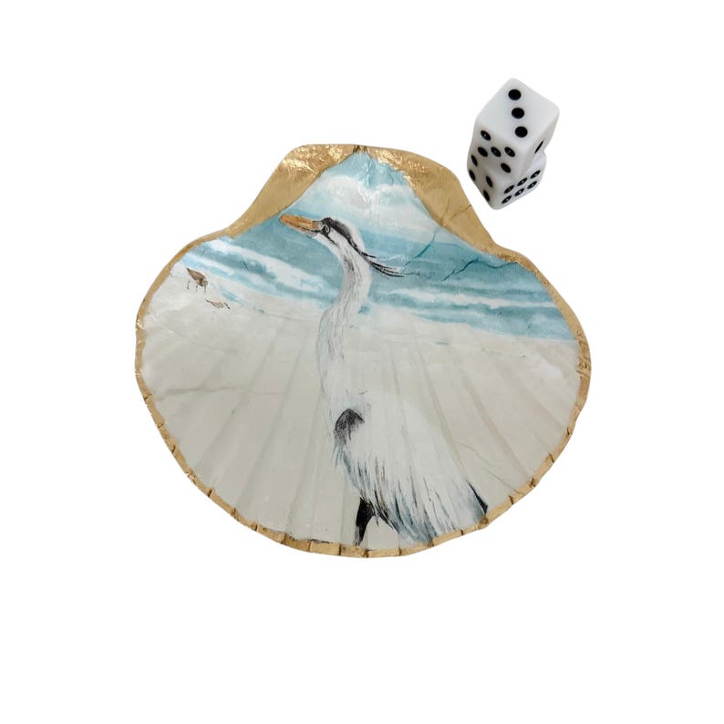 May include: A decorative seashell with a painted heron scene, featuring a white bird on a sandy beach with a blue ocean backdrop. The shell has a gold-painted edge. Two white dice with black dots are next to the shell.