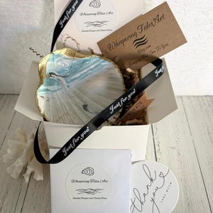 May include: A hand-painted seashell trinket dish with gold trim, presented in a white gift box with a black ribbon that says "Just for you." The dish features a coastal scene in shades of blue and white. The box includes a card that says "Whispering Tides Art."