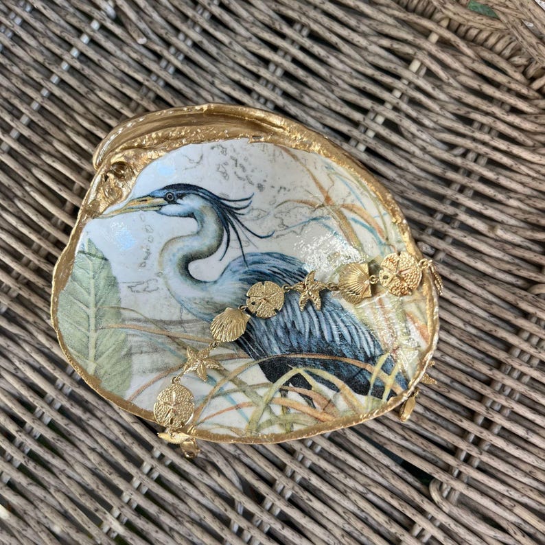 Pu&ograve; includere: Un piatto decorativo a forma di conchiglia con un'illustrazione dipinta di un airone azzurro e bordi dorati. Un braccialetto a catena color oro con piccoli ciondoli a forma di stella marina, dollari della sabbia e conchiglie &egrave; appoggiato all'interno.