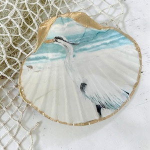A scallop seashell is shown with a white egret coastal bird design. The shell is trimmed in shimmering gold. A coastal decor accent. The shell is resting upon a light tan nautical netting.