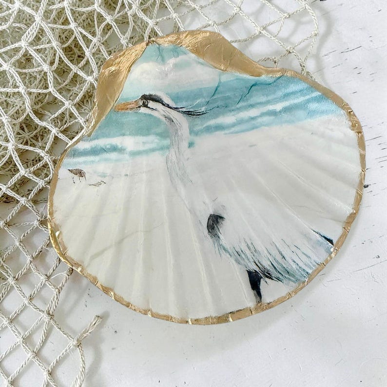 A scallop seashell is shown with a white egret coastal bird design. The shell is trimmed in shimmering gold. A coastal decor accent. The shell is resting upon a light tan nautical netting.