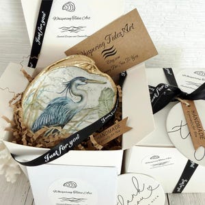 Pu&ograve; includere: Un guscio di ostrica decorativo con un airone blu dipinto a mano e bordi dorati, inserito in una scatola regalo bianca con carta di riempimento marrone. La confezione include un nastro nero con la scritta Just for you e cartellini di Whispering Tides Art.