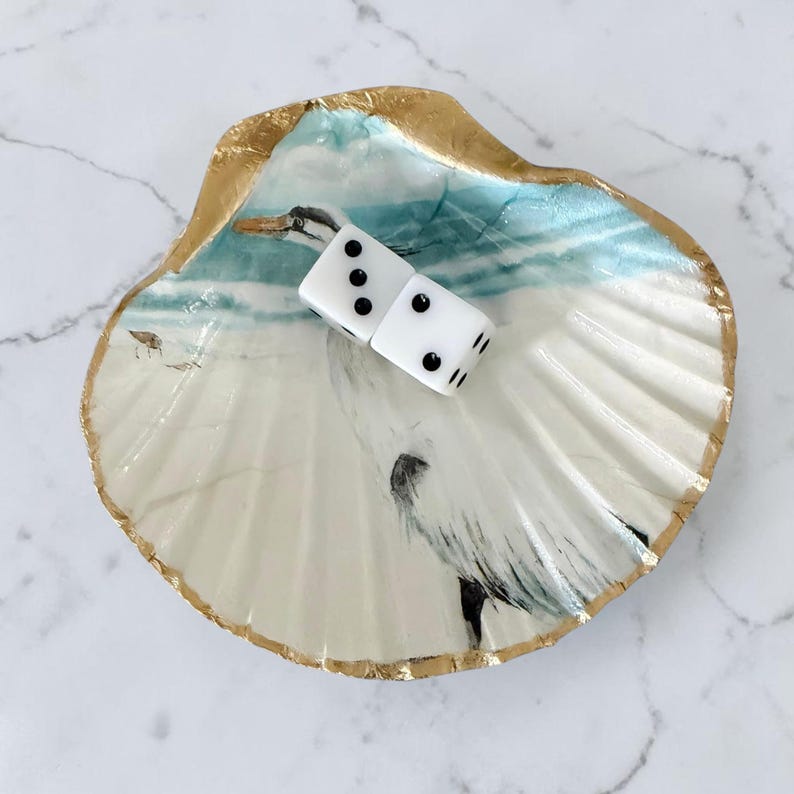 A scallop seashell dice holder for Mahjong in a egret bird design with a beach ocean background. Two dice sit on the shell with the numbers 2 and 3 shown. The shell trim is painted in shimmering gold paint.