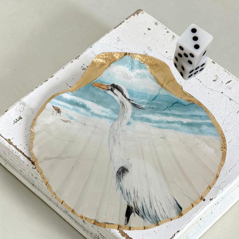 A scallop shell with a white egret design and trimmed in gold edging . Mahjong dice are placed on the right side of the shell with the number three showing.