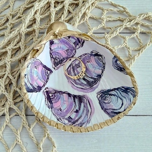 May include: A decorative seashell trinket dish with a gold rim and oyster illustrations in shades of purple, pink, and blue. A gold ring rests inside the dish, which is set against a neutral background.