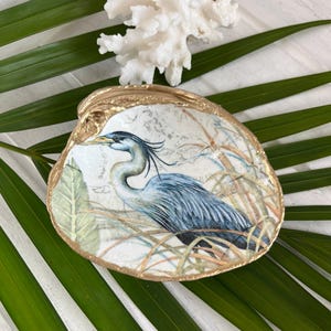 Pu&ograve; includere: Un piatto decorativo a forma di conchiglia con un'illustrazione dettagliata di un airone blu e grigio tra erbe alte. La conchiglia presenta una superficie interna bianca liscia e un bordo dipinto in oro metallizzato, dal look costiero.