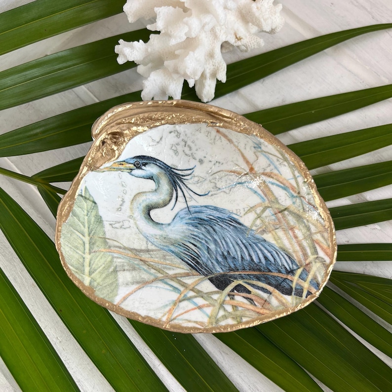 Pu&ograve; includere: Un piatto decorativo a forma di conchiglia con un'illustrazione dettagliata di un airone blu e grigio tra erbe alte. La conchiglia presenta una superficie interna bianca liscia e un bordo dipinto in oro metallizzato, dal look costiero.