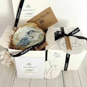 Pu&ograve; includere: Una conchiglia decorativa con dipinto un airone azzurro, inserita in una scatola regalo bianca con carta marrone stropicciata. La scatola include un nastro nero con il testo Just for you, un'etichetta di ringraziamento e una carta Whispering Tides Art.