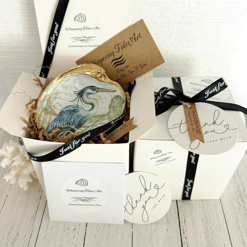 Pu&ograve; includere: Una conchiglia decorativa con dipinto un airone azzurro, inserita in una scatola regalo bianca con carta marrone stropicciata. La scatola include un nastro nero con il testo Just for you, un'etichetta di ringraziamento e una carta Whispering Tides Art.