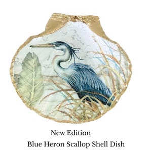 Pu&ograve; includere: Un piatto decorativo a forma di conchiglia di capasanta con un'illustrazione di un airone blu tra canne e fogliame. La conchiglia ha un interno bianco con un bordo dorato. Il testo sottostante recita: New Edition Blue Heron Scallop Shell Dish.