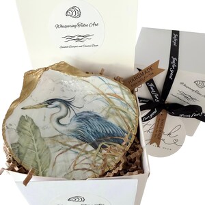 Pu&ograve; includere: Un piatto decorativo a forma di conchiglia di ostrica con un airone blu dipinto tra le canne e un bordo dorato. La conchiglia &egrave; inserita in una scatola regalo bianca con carta riempitiva, un nastro nero "Just for you" e un'etichetta "Handmade".