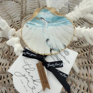 May include: Hand-painted seashell art featuring a white heron against a blue and white beach scene. The shell has a gold-painted edge. Includes a black ribbon with "Just for you" text, a "Thank you" card, and a "HANDMADE" tag.