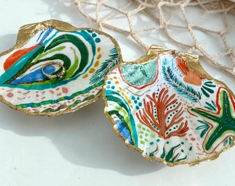 Può includere: Due conchiglie decorative con bordi dipinti in oro, caratterizzate da motivi astratti colorati dipinti a mano, inclusi disegni in verde, arancione e blu. Un piccolo anello d'argento è appoggiato all'interno di una delle conchiglie.