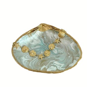 May include: A large, iridescent seashell-shaped dish with a gold-colored rim. A gold-colored necklace with seashell and starfish charms rests inside the dish. The dish has a marbled blue and white interior.
