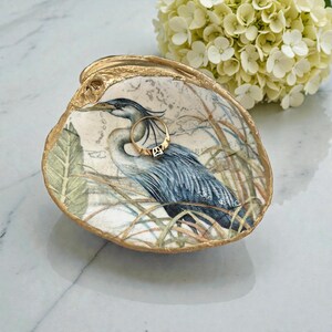 Pu&ograve; includere: Un piattino decorativo a forma di conchiglia di ostrica con un'illustrazione dettagliata di un airone azzurro tra l'erba alta. La conchiglia presenta un bordo dipinto in oro metallizzato e contiene un sottile anello dorato con una pietra chiara.
