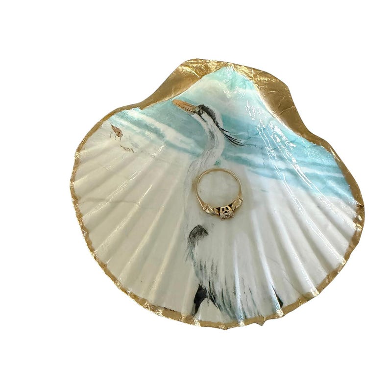 Coastal seashell ring holder with a white egret design. The scallop shell is trimmed in gold edging. A gold diamond wedding ring sits in the middle of the shell.
