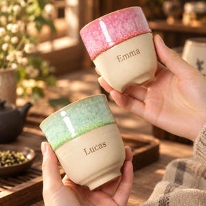 May include: Two ceramic cups with a matte beige base and a glazed rim. One cup features a pink speckled glaze with the name Emma engraved, while the other has a light green speckled glaze with the name Lucas engraved on the side.