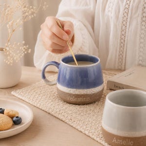 May include: A ceramic mug with a blue glazed top and a speckled white band transitioning into a brown base, featuring the name Valentino engraved on the side. A second mug with a grey top and brown base displays the name Amelie in the foreground.