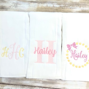May include: Three white baby blankets with embroidered designs. One blanket features the initials "H C" in yellow and pink. Another has the name "Hailey" in pink. The third blanket displays "Hailey" within a circle of yellow dots and a pink bow.