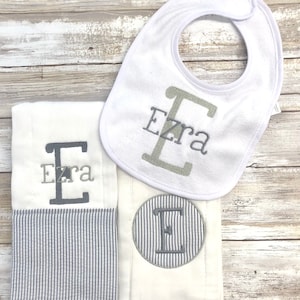 May include: A set of three white baby items with gray embroidery. The items include a bib, a burp cloth, and a washcloth. The bib has the name "Ezra" embroidered on it. The burp cloth has a striped border and the name "Ezra" embroidered on it. The washcloth has a circle with the letter "E" embroidered on it.