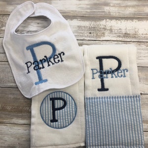 May include: A set of three white baby items with blue and navy blue embroidery. A bib with the name "Parker" embroidered on it, a burp cloth with a blue and white striped circle with the letter "P" embroidered on it, and a burp cloth with the name "Parker" embroidered on it.