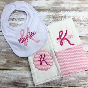 May include: A set of two white baby burp cloths with pink and white stripes. One burp cloth has a pink monogrammed "K" in a circle. The other burp cloth has the name "Kaylee" embroidered in pink with a pink ribbon design. A white bib with the name "Kaylee" embroidered in pink with a pink ribbon design is also included.