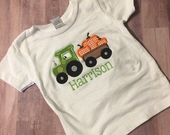 May include: White short-sleeved t-shirt with a green tractor pulling a wagon full of orange pumpkins. The name "Harrison" is embroidered in green below the tractor.
