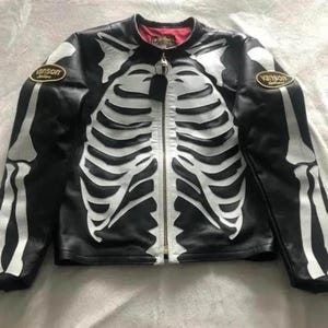 May include: Black leather jacket with a white skeleton design. The jacket features a zipper closure and two patches with the word "Vanson". The interior lining is red.