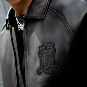 Puede incluir: Chaqueta de cuero negra con cuello y cierre de cremallera. La chaqueta presenta un parche negro en el pecho con el texto "AVIREX ESTABLISHED 1975". Dise&ntilde;o cl&aacute;sico.