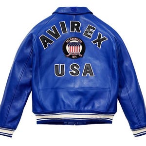 K&ouml;nnte beinhalten: K&ouml;nigsblaue Lederjacke mit dem Wort "AVIREX" in wei&szlig;en Buchstaben &uuml;ber den Schultern. Darunter befindet sich ein kreisf&ouml;rmiger Aufn&auml;her mit der Aufschrift "ALL AMERICANS ESTABLISHED 1975 USA". Die Jacke hat einen wei&szlig;-blauen Streifen.