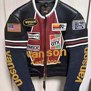 Puede incluir: Chaqueta de cuero negra, roja y azul marino con múltiples parches, incluyendo una bandera americana y logotipos de "Vanson Leathers". La chaqueta tiene cremallera frontal y rayas blancas y rojas.