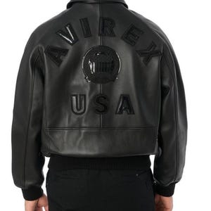 Puede incluir: Chaqueta bomber de cuero negro con la palabra "AVIREX" arqueada en la espalda, encima de un emblema circular y la palabra "USA". La chaqueta tiene dobladillo y pu&ntilde;os de canal&eacute;.