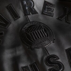 Puede incluir: Primer plano de una chaqueta de cuero negro con letras en relieve y brillantes que deletrean "AIREX" y otras letras. Un parche circular presenta un emblema con el texto "ALL AMERICAN" y "ESTABLISHED 1975". La chaqueta tiene una superficie texturizada.