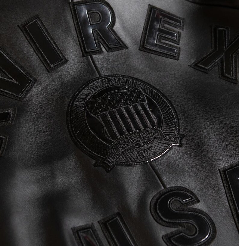 Puede incluir: Primer plano de una chaqueta de cuero negro con letras en relieve y brillantes que deletrean "AIREX" y otras letras. Un parche circular presenta un emblema con el texto "ALL AMERICAN" y "ESTABLISHED 1975". La chaqueta tiene una superficie texturizada.