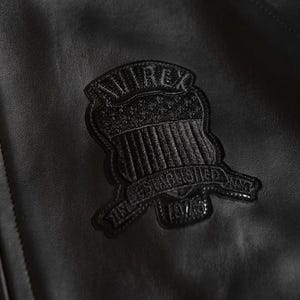 Puede incluir: Primer plano de una chaqueta de cuero negra con un parche texturizado. El parche tiene forma de escudo, con la palabra "AVIREX" en la parte superior y "ESTABLISHED NYC 1975" en la inferior. La chaqueta tiene un acabado liso y mate.
