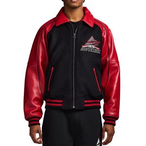 May include: A black and red varsity jacket featuring a full-length front zipper, a pointed collar, and long red sleeves. The chest displays an embroidered logo with the text 1975 AVIREX. The jacket includes ribbed cuffs and a ribbed hem in black and red.