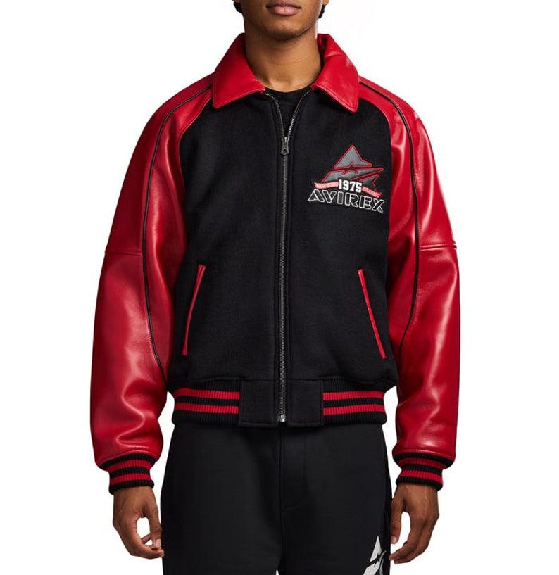 May include: A black and red varsity jacket featuring a full-length front zipper, a pointed collar, and long red sleeves. The chest displays an embroidered logo with the text 1975 AVIREX. The jacket includes ribbed cuffs and a ribbed hem in black and red.